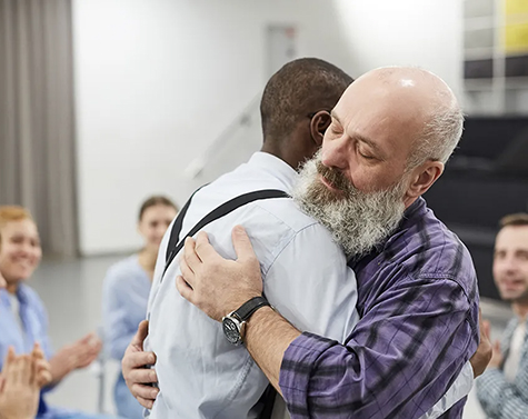 Two men, one white with a beard the other African American, embrace during a group support meeting.